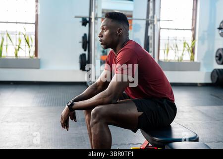 African American man resting on weight bench in gym wearing red athletic T-shirt, smartwatch. Fitness, strength, motivation, training, sport, activewe Stock Photo
