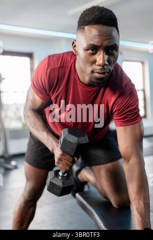 African American man in red T-shirt doing one-arm hexagonal dumbbell row on flat bench at gym. Strength, fitness, training, sport, motion, discipline, Stock Photo