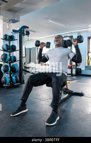 Senior African American man lifting dumbbells on bench in fitness studio with medicine ball rack. Fitness, strength, health, endurance, motivation, we Stock Photo