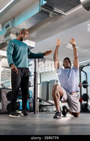 Senior African American male trainer guiding client doing forward lunge at gym with kettlebell rack. Fitness, coaching, strength, motivation, workout, Stock Photo