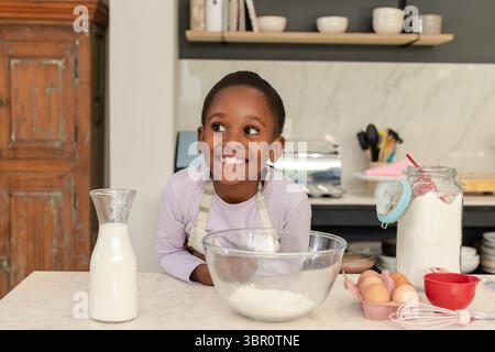 Smiling African American girl in apron mixing flour and milk in glass bowl at home kitchen. Children, culinary, rustic, home cooking, cheerful, domest Stock Photo