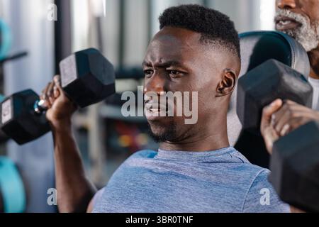 African American man pressing hex dumbbells on gym bench with senior trainer assisting. Fitness, strength, coaching, training, determination, well-bei Stock Photo