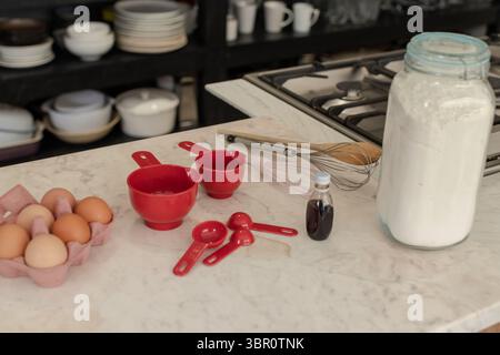 Red measuring cups and spoons are sitting on marble countertop beside egg carton, whisks, stovetop. Cooking, baking, kitchenware, culinary, culinary t Stock Photo
