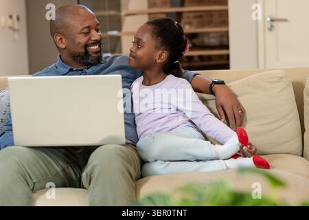 African American father and daughter sitting on beige couch at home using laptop near green plant. Family bonds, nurturing, comfort, technology, home Stock Photo