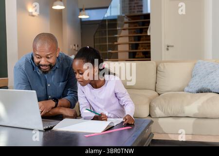 African American father and daughter using laptop writing notebook holding pencil in living room. Family, education, technology, interior, bonding, mo Stock Photo
