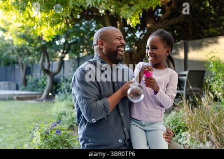 African American father and daughter holding pink bubble wand in garden blowing soap bubbles. Family, outdoor, playful, nature, joyful, leisure, conne Stock Photo