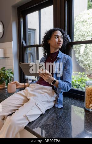 Non-binary person sitting on granite counter in home kitchen holding tablet and mug by potted plant. Contemporary, lifestyle, minimalism, wellness, in Stock Photo