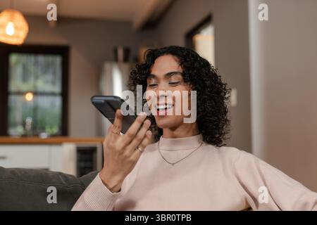 Non-binary person reclining on gray sofa in living room holding smartphone by countertop speaking. Lifestyle, modern, cozy, minimalist, technology, re Stock Photo