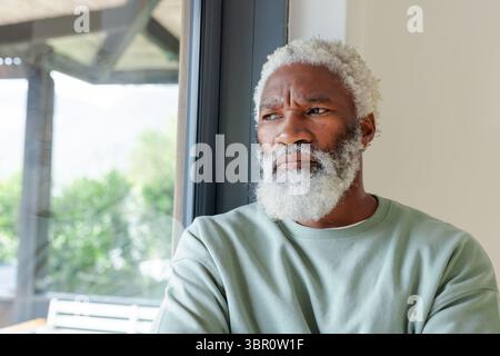 Senior African American man standing in sunroom looking out glass door at wooden bench, copy space. Contemplation, serenity, relaxation, leisure, natu Stock Photo
