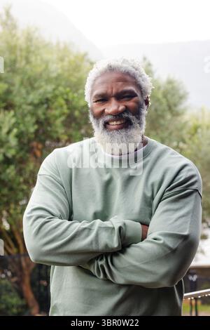 Senior African American man standing with arms crossed wearing light green sweatshirt in backyard. Portrait, wellness, nature, casual, tranquility, li Stock Photo