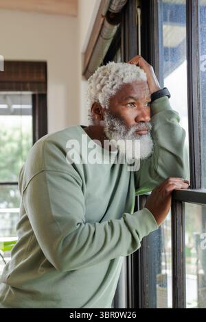 Senior African American man leaning on window sill at home, wearing fitness tracker, copy space. Contemplation, relaxation, mindfulness, cozy, domesti Stock Photo