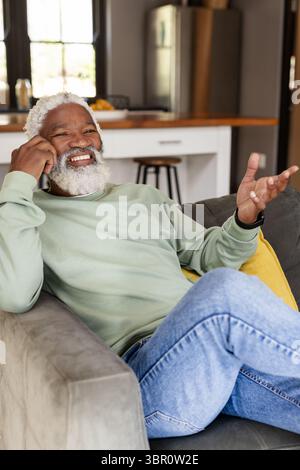 Mature African American man talking on smartphone while sitting on gray sofa in open living area. Casual, modern, cozy, contemporary, home, lifestyle, Stock Photo
