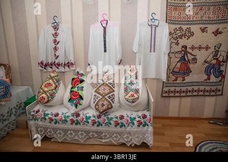 Vibrant embroidered pillows with floral and geometric motifs on a woven bench beneath hanging vyshyvanka shirts and a folk tapestry at a cultural exhi Stock Photo