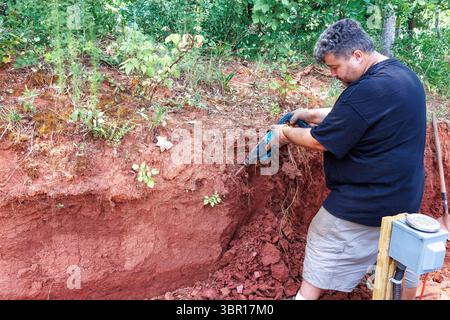 Person uses jackhammer tool to dig into red soil, revealing layer patterns roots in yard full Stock Photo