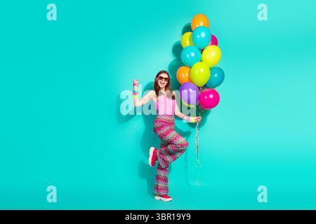 Smiling woman holding colorful balloons on turquoise background, wearing retro hippie fashion, expressing carefree joy Stock Photo