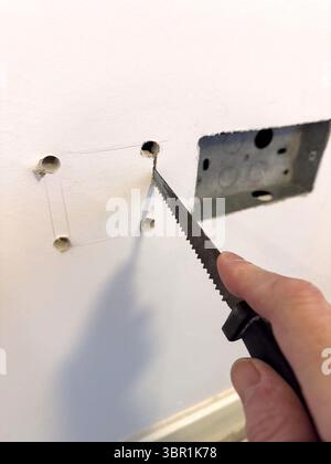 Hand using a drywall plasterboard saw to cut a square hole in a white wall to install an electrical outlet box or pattress uk Stock Photo