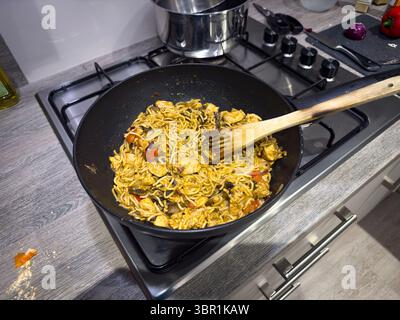 A wok full of stir-fried noodles with chicken and vegetables sits on a modern kitchen stove with a wooden spoon  asian cuisine Stock Photo