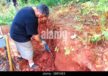 Contractor uses jackhammer to break up red soil for construction project in forested area on works day. Stock Photo