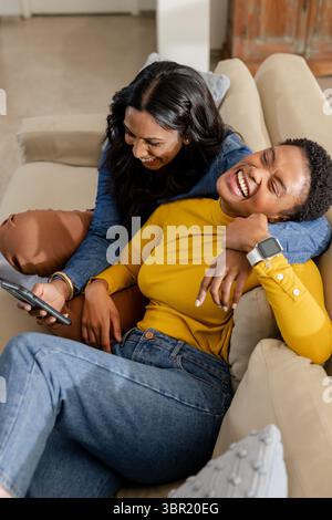 Diverse female friends laughing and using smartphone while lounging on beige couch in living room. Friendship, camaraderie, relaxation, home, casual, Stock Photo
