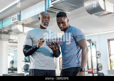 African American men studying workout data on tablet at fitness center with dumbbell rack and bench. Personal training, gym, strength, motivation, wel Stock Photo