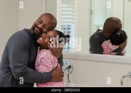 African American father and daughter wearing bathrobe hugging and smiling by sink in bathroom. Family, caregiving, intimacy, tenderness, domestic, aff Stock Photo