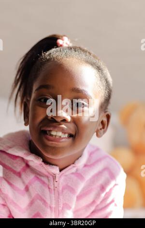 African American girl sitting at home wearing pink hoodie smiling at camera with teddy bear nearby. Children, childhood, comfort, innocence, warmth, r Stock Photo