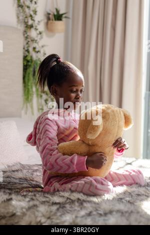 African American girl in pink hooded onesie hugging plush teddy bear on bed near potted plant. Children, bedroom, cozy, comfort, relaxation, home deco Stock Photo
