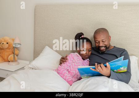African American father reading children's book to daughter in bedroom on bed with teddy bear. Family, bonding, education, cozy, warm, storytelling, c Stock Photo