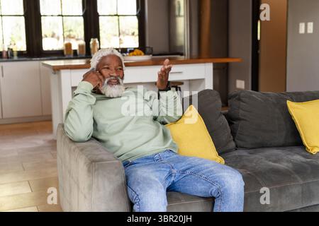 Senior African American man lying on gray sofa in sweatshirt, talking on smartphone in living room. Comfort, relaxation, domestic, contemporary, leisu Stock Photo