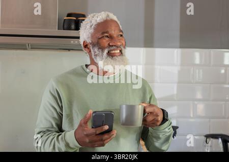 Senior African American man using smartphone with smartwatch and sipping from gray mug in kitchen. Aging, technology, relaxation, lifestyle, modern, h Stock Photo