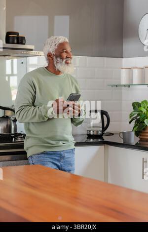 Senior African American man leaning against counter in kitchen holding smartphone smiling. Contemporary, lifestyle, minimalist, technology, domestic, Stock Photo