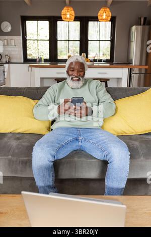 Senior African American man smiling and tapping smartphone on gray sofa at home with open laptop. Elderly, relaxation, modern, connectivity, comfort, Stock Photo