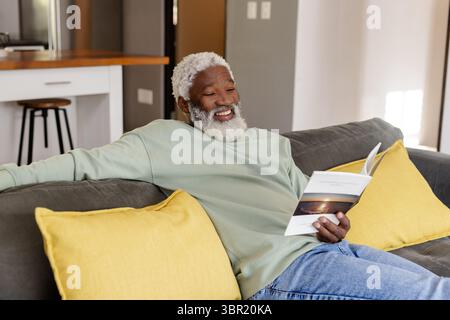 Senior African American man sitting on sofa with yellow pillows reading book next to kitchen island. Comfortable, relaxation, cozy, contemporary, life Stock Photo