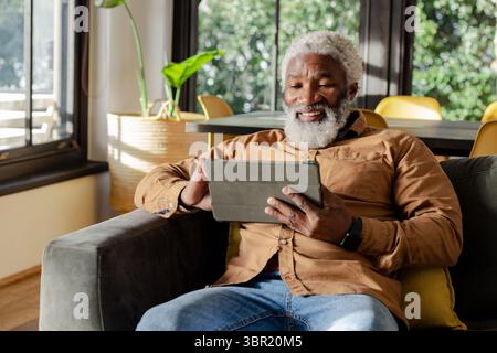 Senior African American man sitting on sofa at home using tablet with green plant beside window. Senior, leisure, technology, relaxation, cozy, modern Stock Photo