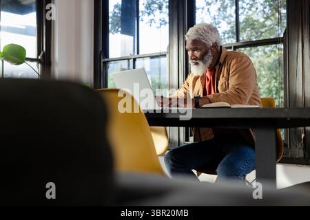 mature African American man typing on laptop at home office table by large windows, copy space. Professional, workspace, modern, entrepreneur, product Stock Photo