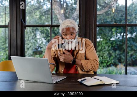 Senior African American man talking on smartphone and gesturing in home office with laptop notebook. Professional, technology, workspace, lifestyle, m Stock Photo
