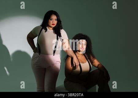 Diverse female friends posing around low block in studio wearing black suspenders with green wall. Minimalist, fashion, contemporary, stylish, express Stock Photo