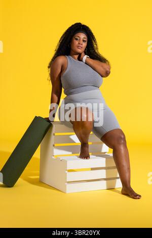 African American woman in grey sportswear sitting on crate in yellow studio holding green yoga mat. Fitness, wellness, activewear, studio, motivationa Stock Photo