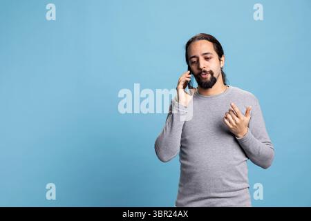 Middle eastern guy chatting on a phone call with a friend while he stands against blue background, having fun on a telephone line in studio. Young casual adult with long hair calling people. Stock Photo