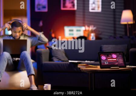 African American man works on laptop at home next to tablet showing alert, notification of a hacking attempt in the cozy living room. Cyberattack and data breach highlight the cyber threat. Stock Photo