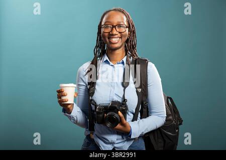 Portrait of professional female photographer, smiling with backpack and coffee cup in hand. Black woman holding dslr and looking directly at camera, ready for new photography session. Stock Photo