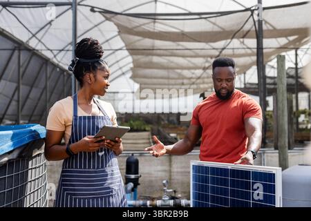 African American colleagues holding tablet and examining solar panel setup inside greenhouse. Agriculture, sustainability, innovation, technology, tea Stock Photo