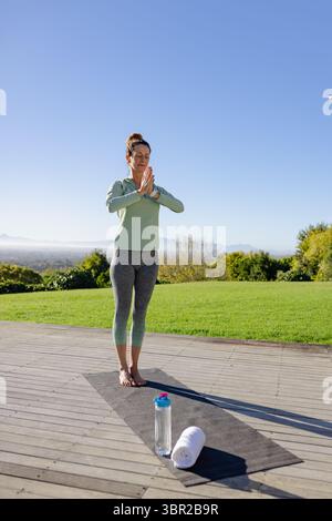 Woman practicing yoga in sportswear on wooden deck by lawn with yoga mat, towel, water bottle. Wellness, tranquility, serenity, outdoor, nature, fitne Stock Photo
