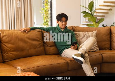 Asian man sitting on sofa at home, checking smartphone near plant, floating staircase, copy space. Modern, casual, cozy, stylish, relaxation, interior Stock Photo