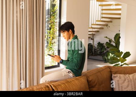 Adult Asian man sitting on couch by beige curtains in living room using smartphone and smartwatch. Modern, interior, technology, lifestyle, casual, co Stock Photo