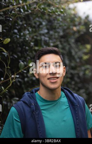 Hispanic man standing on garden pathway smiling at camera wearing teal tee and navy hoodie. Portrait, nature, outdoor, casual, greenery, wellness, lif Stock Photo