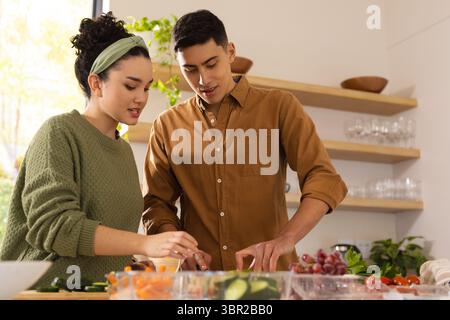 Diverse couple slicing fresh produce on cutting board in modern kitchen with clear glass bowls. Group, culinary, contemporary, healthy living, lifesty Stock Photo
