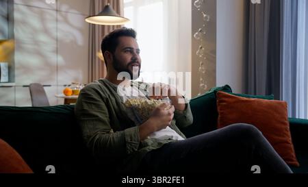 Caucasian bearded joyful man business male businessman guy homeowner buyer client customer falling on sofa couch relaxing eating popcorn unhealthy Stock Photo