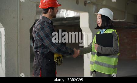 Two Caucasian Japanese builders contractors man woman industry workers hardhat engineers team cooperation shaking hands handshake gesture agreement Stock Photo