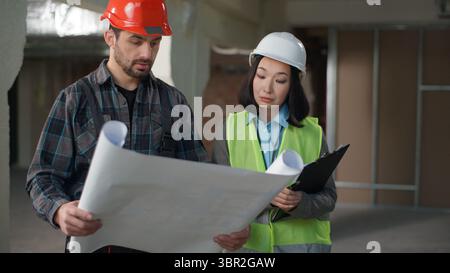 Multiethnic builders in uniform discussing blueprint inspection business engineers Caucasian man Korean woman hardhat discuss plan construction Stock Photo
